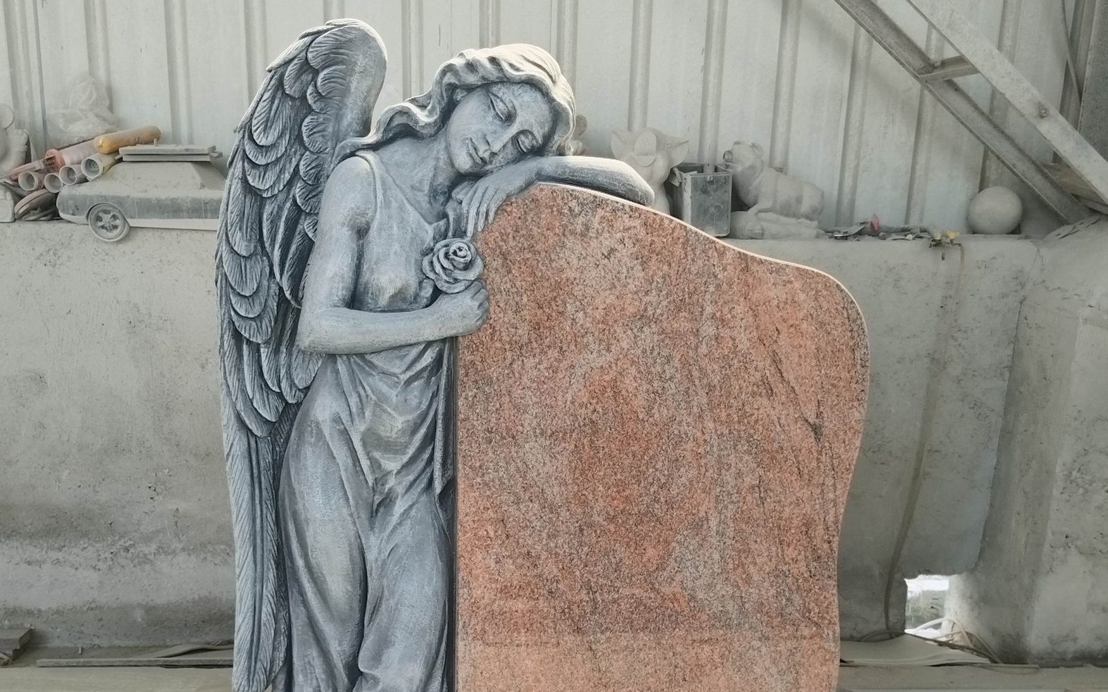 Grabstein aus rötlichem Granit mit gemeißeltem Engel, der eine Rose hält und sich an den Stein lehnt, in der Werkstatt