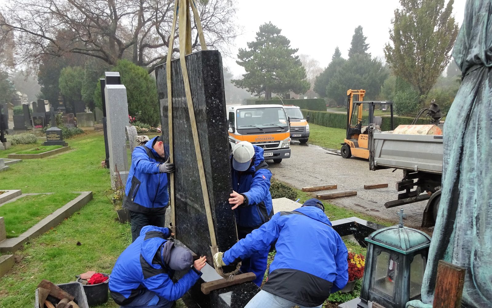 Mitarbeiter von Steinmetz Stockerer richten einen Grabstein aus schwarzem Granit mit Hebegurten auf und setzen ihn standsicher auf den Sockel
