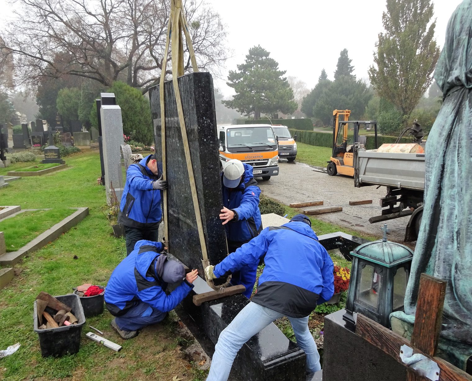 Vier Mitarbeiter von Steinmetz Stockerer setzen auf einem Wiener Friedhof einen Grabstein aus schwarzem Granit mit Hebegurten und Kran standsicher auf den Sockel